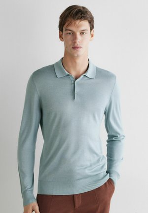 Junger Mann trägt ein hellblaues langärmeliges Poloshirt und braune Hose, steht mit einer Hand in der Tasche vor einer weißen Wand.