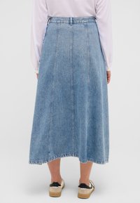 Jeansrock in Hellblau, knielang, mit einem taillierten Bund und sichtbaren Nahtdetails. Kombiniert mit beigen Schuhen mit schwarzen Akzenten.