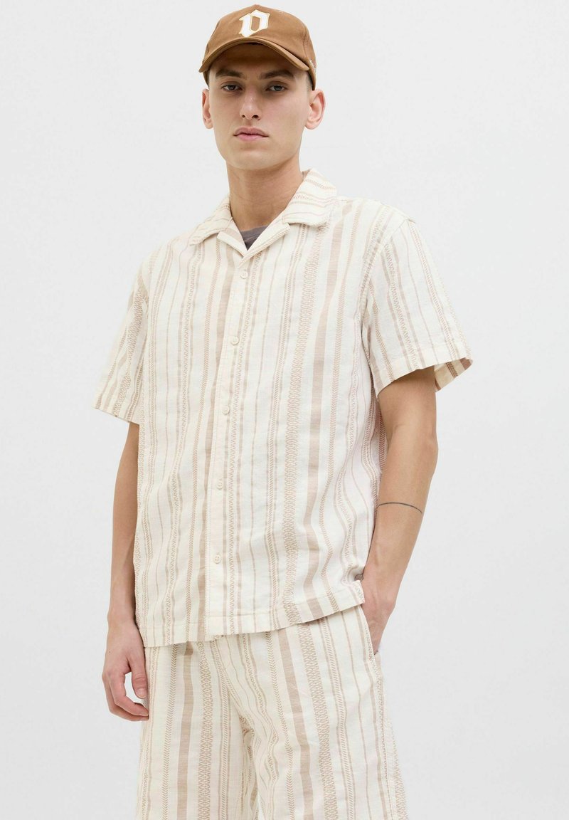 Chemise à manches courtes, à boutons, en tissu léger avec des rayures verticales beige et crème. Présente un col camp et une coupe décontractée.