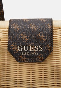 Sac en simili cuir marron à motif avec une base en paille tressée. Doté d'un logo métallique "GUESS" et de l'insigne "EST 1981" sur un rabat rectangulaire.