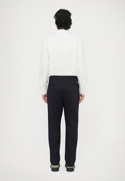 Biela košeľa s dlhými rukávmi a rovný chrbtom, v kombinácii s námorníckymi nohavicami, ktoré majú hladkú textúru a jemné detaily na vreckách. Čierne topánky dopĺňajú vzhľad.