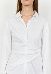 Camicia bianca a maniche lunghe con colletto, caratterizzata da un design avvolgente e una texture liscia. La parte frontale presenta pieghe sottili per un tocco di dettaglio.