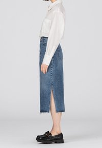 Kvinna som står på sidan i en vit skjorta med knappar, hög midja denimkjol med slits och svarta loafers mot en neutral bakgrund.