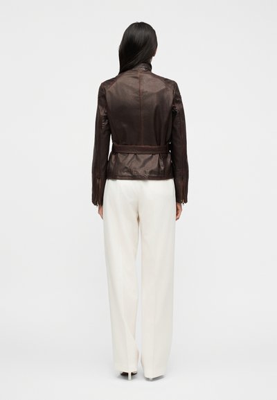 Γυναίκα που στέκεται γυρισμένη πλάτη, φοράει εφαρμοστή σκούρα καφέ δερμάτινη ζακέτα με ζώ νη και λευκό παντελόνι με φαρδιά πατζάκια, σε λευκό φόντο.