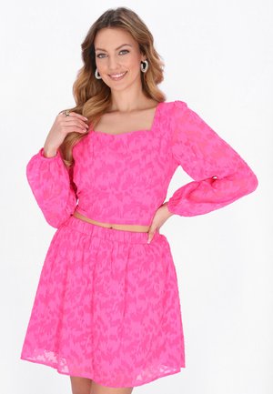 Usměvavá žena s vlnitými vlasy, která nosí kruhové náušnice a výrazný růžový dvoudílný outfit s dlouhými rukávy a vzorovanou sukní.