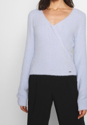 Femme portant un pull cache-cœur côtelé bleu clair à manches longues et un pantalon noir, debout devant un fond uni.