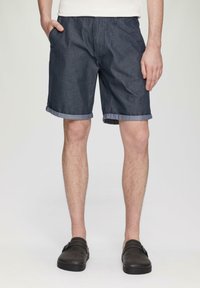 Shorts en denim bleu foncé avec des ourlets retroussés, dotés d'un devant plat et de poches latérales, associés à des chaussures à enfiler sombres.