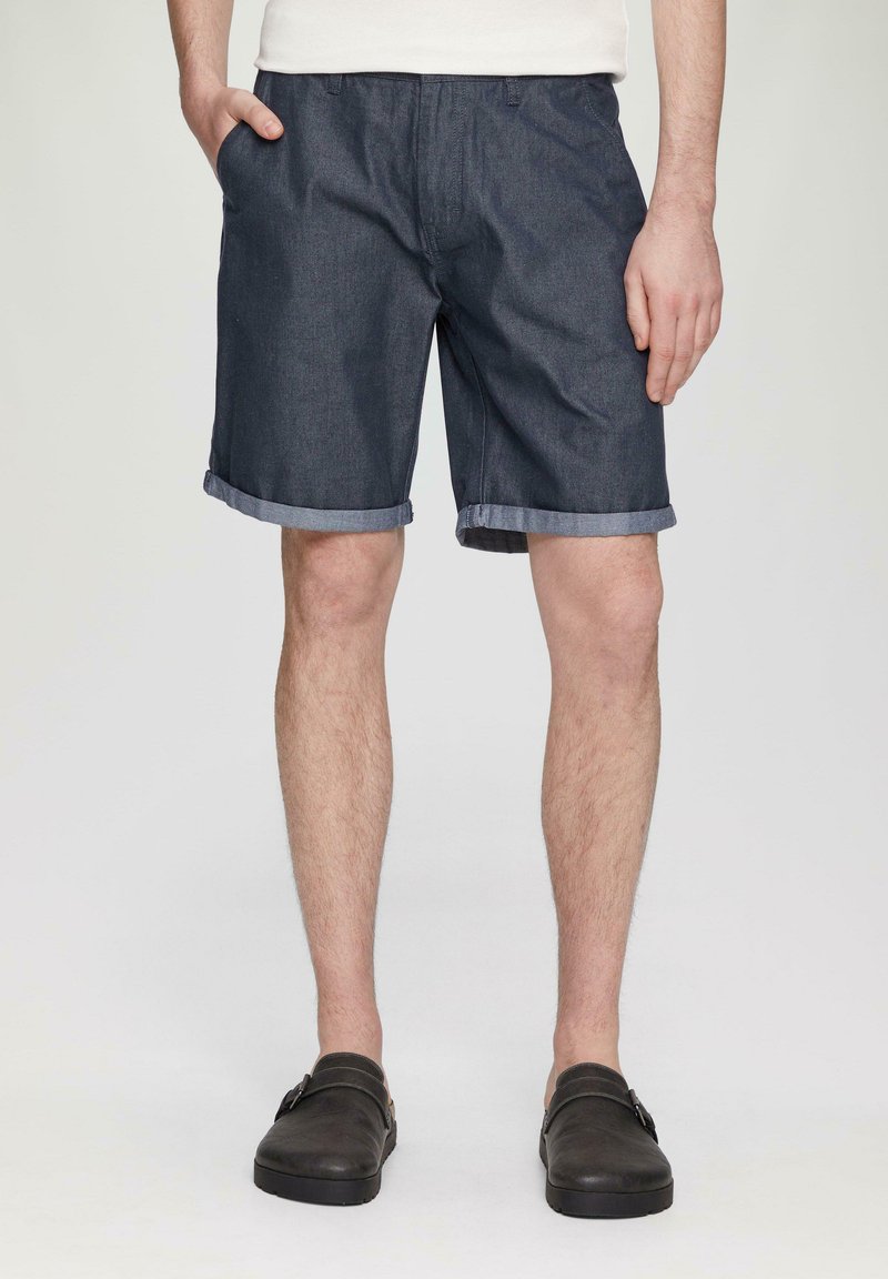 Shorts en denim bleu foncé avec des ourlets retroussés, dotés d'un devant plat et de poches latérales, associés à des chaussures à enfiler sombres.