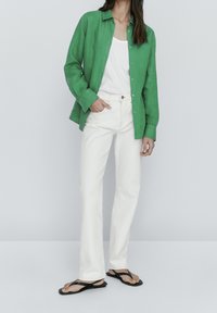 Woman wearing a bright green button-up shirt, white sleeveless top, off-white straight-leg jeans, and black flat sandals standing against a plain background.