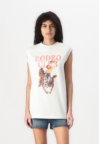 Bala bez piedurknēm T-krekls ar grafisku attēlu no rodeo ainas. Teksts "RODEO KLUBS" ir oranžs. Saskaņots ar džinsu šortiem.