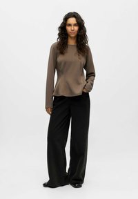 Rjav dolg rokav z oprijetim telesom in širokim dnom; v kombinaciji z oversized črnimi širokimi hlačami in svetlečimi črnimi čevlji.