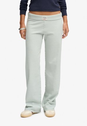 Kvinde iført lysegrå ribstrikkede vidde bukser og beige sneakers, med en navy crop top med lange ærmer og sølvarmbånd på venstre håndled.