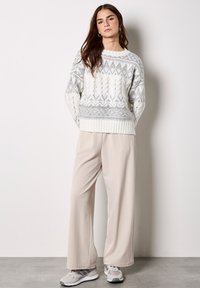 Gestrickter cremefarbener Pullover mit grauen geometrischen Mustern, kombiniert mit beigefarbenen weiten Hosen und grauen Turnschuhen. Weiche Texturen und entspannte Passform.