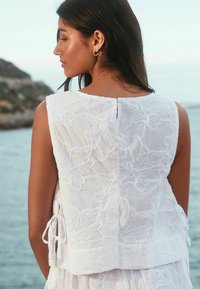 Blusa senza maniche bianca ricamata con motivi floreali, dotata di chiusura a bottone sul retro e lacci ai lati. Tessuto strutturato.