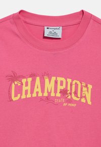 Ροζ βαμβακερό t-shirt με κίτρινο κείμενο «CHAMPION» και γραφικά με φοίνικες. Στρογγυλή λαιμόκοψη με κοντά μανίκια, κανονική εφαρμογή.