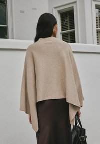 Poncho de lana beige con un diseño suelto y drapeado. Cuello redondeado, dobladillo asimétrico, combinado con una falda marrón oscuro y un bolso tote negro.