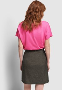 Mujer con pelo rojo hasta los hombros, vestida con una camiseta rosa brillante de Manga corta y una falda oscura hasta la rodilla, de pie contra un fondo claro.