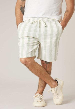 Man die witte en lichtgroene verticale gestreepte shorts draagt, witte sneakers, wit T-shirt, staat met gekruiste benen en handen in de zakken.