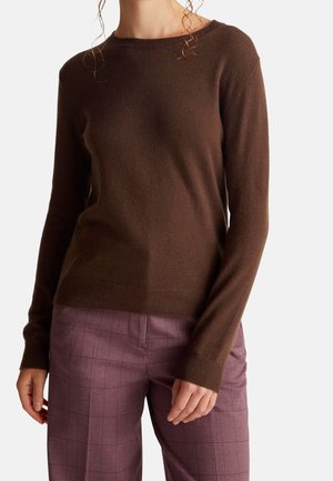 Femme portant un pull marron à manches longues et un pantalon à carreaux violet, debout avec les bras détendus devant un fond uni.