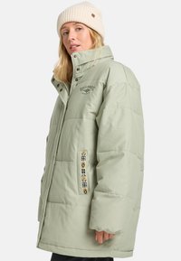 Frau in einer hellgrünen gepolsterten Jacke mit gesticktem Logo und gemusterten Taschendetails, kombiniert mit einer cremefarbenen Strickmütze.