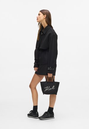 Fekete dzsekit, szoknyát és tornacipőt viselő nő, aki egy fekete táskával áll, amelyen a "Karl" logó látható, profilból, fehér háttér előtt.