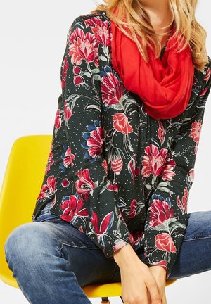Vrouw die een donker bloemig blouse, een rode sjaal en blauwe jeans draagt, zittend met gekruiste benen op een gele stoel tegen een witte achtergrond.