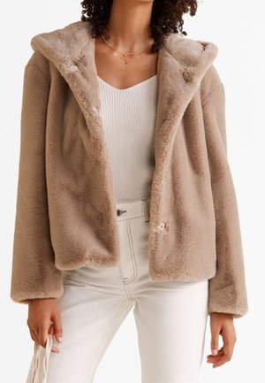 Frau trägt eine beige Kapuzenjacke aus Kunstfell über einem weißen, gerippten Top und einer weißen High-Waist-Hose und hält eine Tasche mit cremefarbenem Riemen.