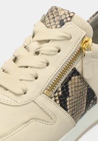 Μπεζ αθλητικό παπούτσι με χρυσή λεπτομέρεια φερμουάρ, με διακοσμητικό σχέδιο φίδι. Υφή δέρματος με επίπεδα κορδόνια και λαστιχένια σόλα.