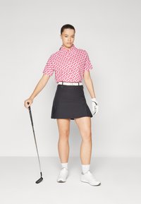 Red and white patterned short-sleeve golf shirt paired with a black pleated skirt, held with a belt. A golf club is held in one hand.