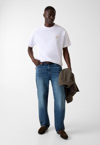 Biała bawełniana koszulka z małym logo, niebieskie dżinsy i brązowe mokasyny. Model trzyma oliwkową kurtkę zarzuconą na jedno ramię.