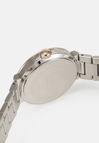 Montre en acier inoxydable avec un bracelet en argent, accents en or rose, et une couronne texturée. Dos lisse avec logo gravé et détails.