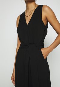 Černý overal bez rukávů má výstřih do V, pásek v oblasti pasu a boční kapsy. Je vyroben z hladkého, splývavého materiálu s volným střihem.