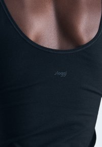 Nærbilde av en mørkhudet person som har på seg en svart ermeløs topp med et "sloggi"-logo på brystet.
