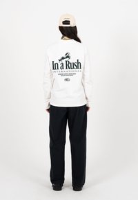 Persona vistiendo una camisa blanca de manga larga con el texto "In a Rush International" y un gráfico de un caballo en la espalda, pantalones negros, gorra beige, de pie mirando hacia atrás.