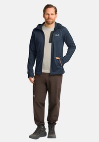 Giacca a hood blu marina con zip frontale, abbinata a una maglietta grigia chiara e pantaloni marroni. Stivali da trekking neri completano l'outfit.