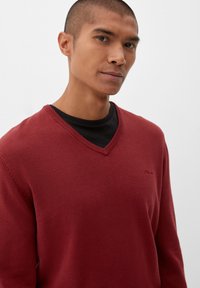 Rode v-hals trui van zachte stof, met ribgebreide manchetten en halslijn, gelaagd over een zwart shirt. Soepele textuur met minimale branding.