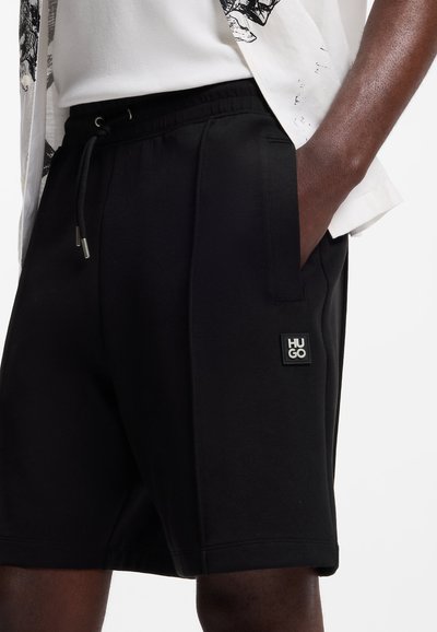 Homme portant un short HUGO noir à cordon de serrage avec poches latérales et une chemise blanche, main dans la poche, vu de la taille jusqu'au milieu des cuisses.