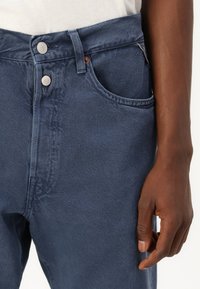 Donkerblauwe denim jeans met een hoge taille, twee voorzakken en zilveren knopen. De stof heeft een gladde textuur met gestikte details.