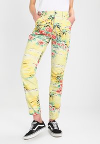 Gula blommönstrade byxor med tropiska motiv som visar rosa blommor och gröna löv; gjorda av mjukt tyg med rak ben-design.