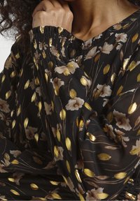 Blusa nera con motivi floreali e pois dorati, caratterizzata da polsini arricciati e una scollatura con bottoni, realizzata in tessuto leggero e trasparente.