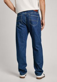 Pantalones vaqueros de mezclilla azul oscuro, corte relajado, mostrando la parte trasera con dos bolsillos traseros y costuras en contraste. Llevados con una camiseta de color claro y zapatos.