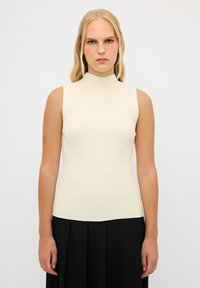 Geribbelde mouwloze top in crème met een hoge kraag, voorzien van kleine decoratieve stippen. Aansluitende silhoutte; zachte textuur. Gecombineerd met een zwarte rok.
