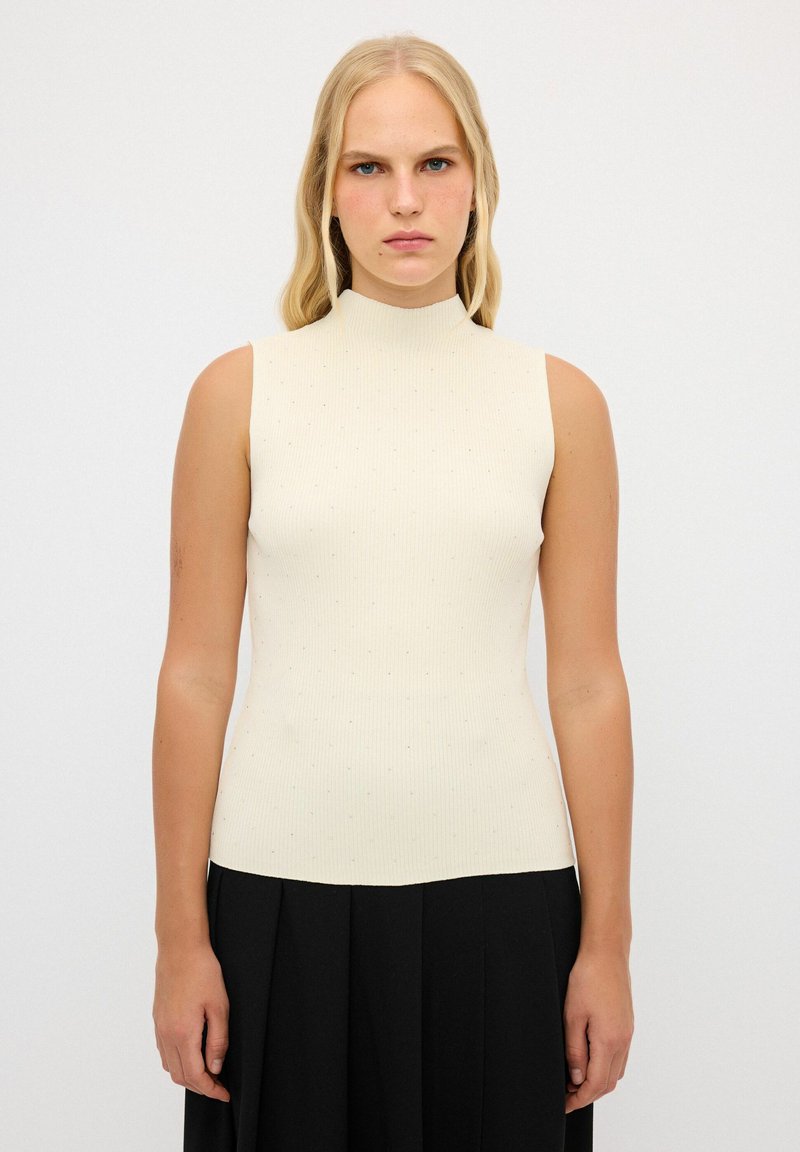 Geribbelde mouwloze top in crème met een hoge kraag, voorzien van kleine decoratieve stippen. Aansluitende silhoutte; zachte textuur. Gecombineerd met een zwarte rok.