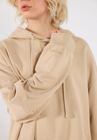 Beige oversized hoodie with a drawstring hood, ribbed cuffs, and subtle "URBAN" embroidery on the sleeve. Soft, smooth fabric texture.
