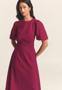 Fuksia mekko, jossa on lyhyet puffihihat, istuva vartalo ja valuva hame. Sisältää hienovaraisia ompeluyksityiskohtia vyötäröllä. Valmistettu kevyestä kankaasta.