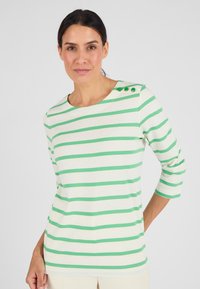 Vrouw in een wit en groen horizontaal gestreept shirt met drie groene knopen op de schouder, staande tegen een effen achtergrond.