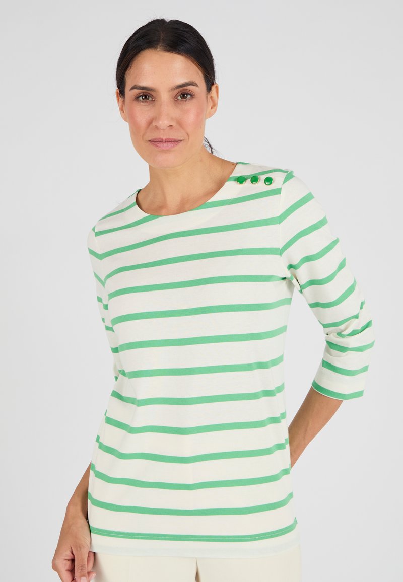 Vrouw in een wit en groen horizontaal gestreept shirt met drie groene knopen op de schouder, staande tegen een effen achtergrond.