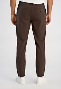 Brązowe spodnie na miarę o wąskim kroju, z dwoma tylnymi kieszeniami i zapięciami na guziki, wykonane z teksturowanej tkaniny, noszone z białymi sneakersami.