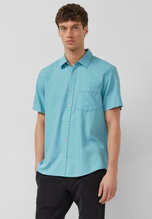 Młody mężczyzna ubrany w jasnoniebieską, krótki rękaw, koszulę na guziki z kieszenią na piersi oraz czarne spodnie, stojący na jasnym, jednolitym tle.