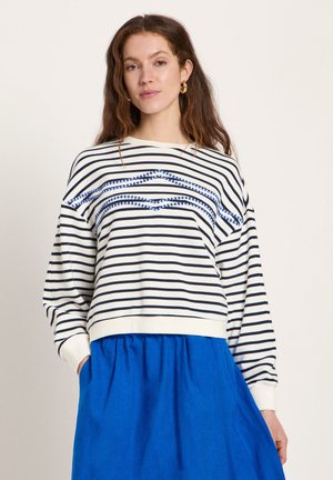 Frau mit langen braunen Haaren, die goldene Creolen, einen weiß-schwarz gestreiften Pullover mit blauem Stickmuster und einen blauen Rock trägt, Hände in den Taschen.